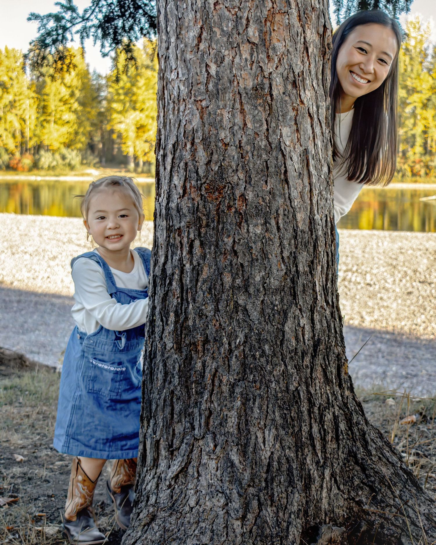 Child in denim overalls plays peek-a-boo behind a large tree trunk in a sunlit forest setting.