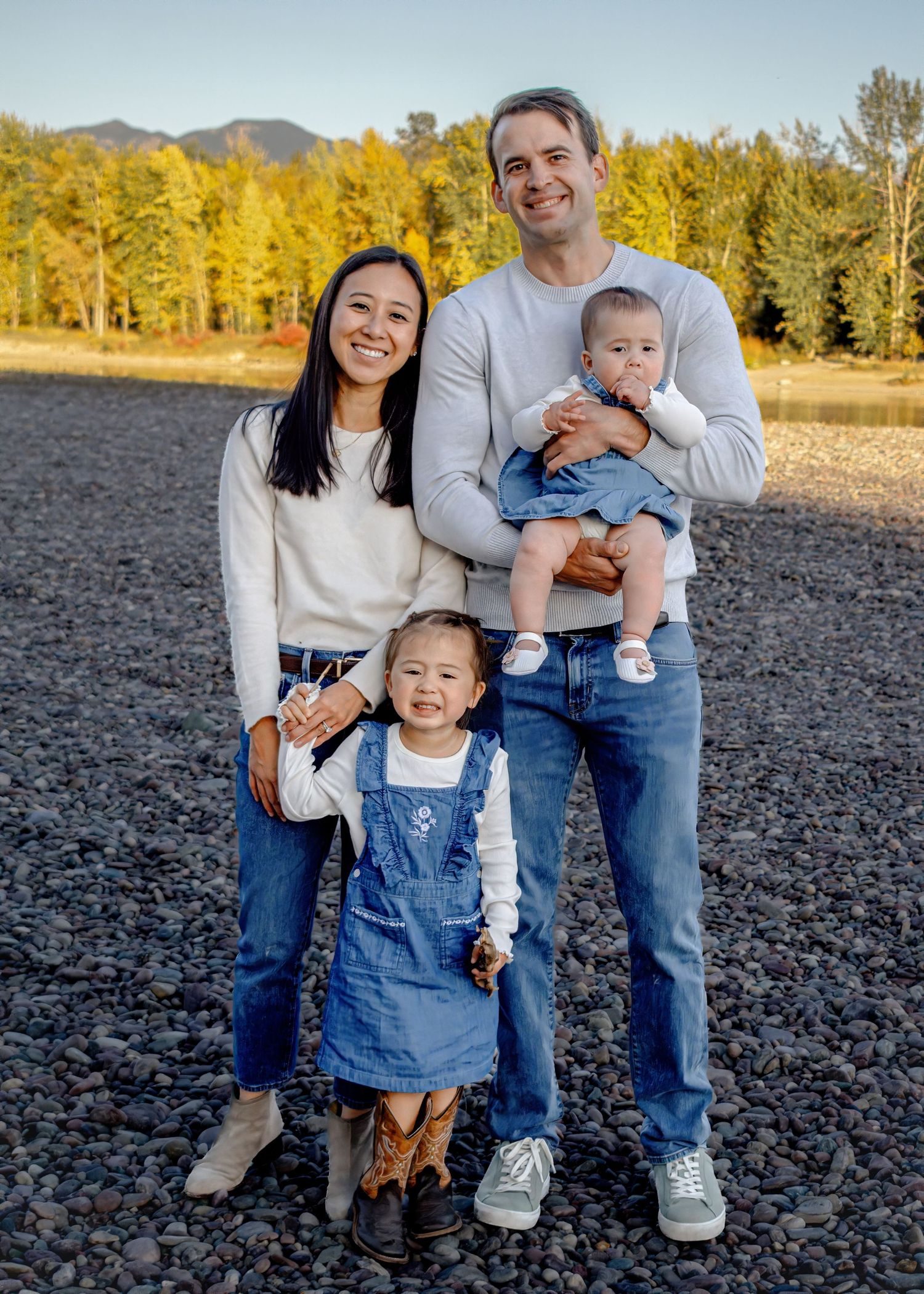 A family in casual attire poses together on dark soil with mountains and autumn trees in the background.