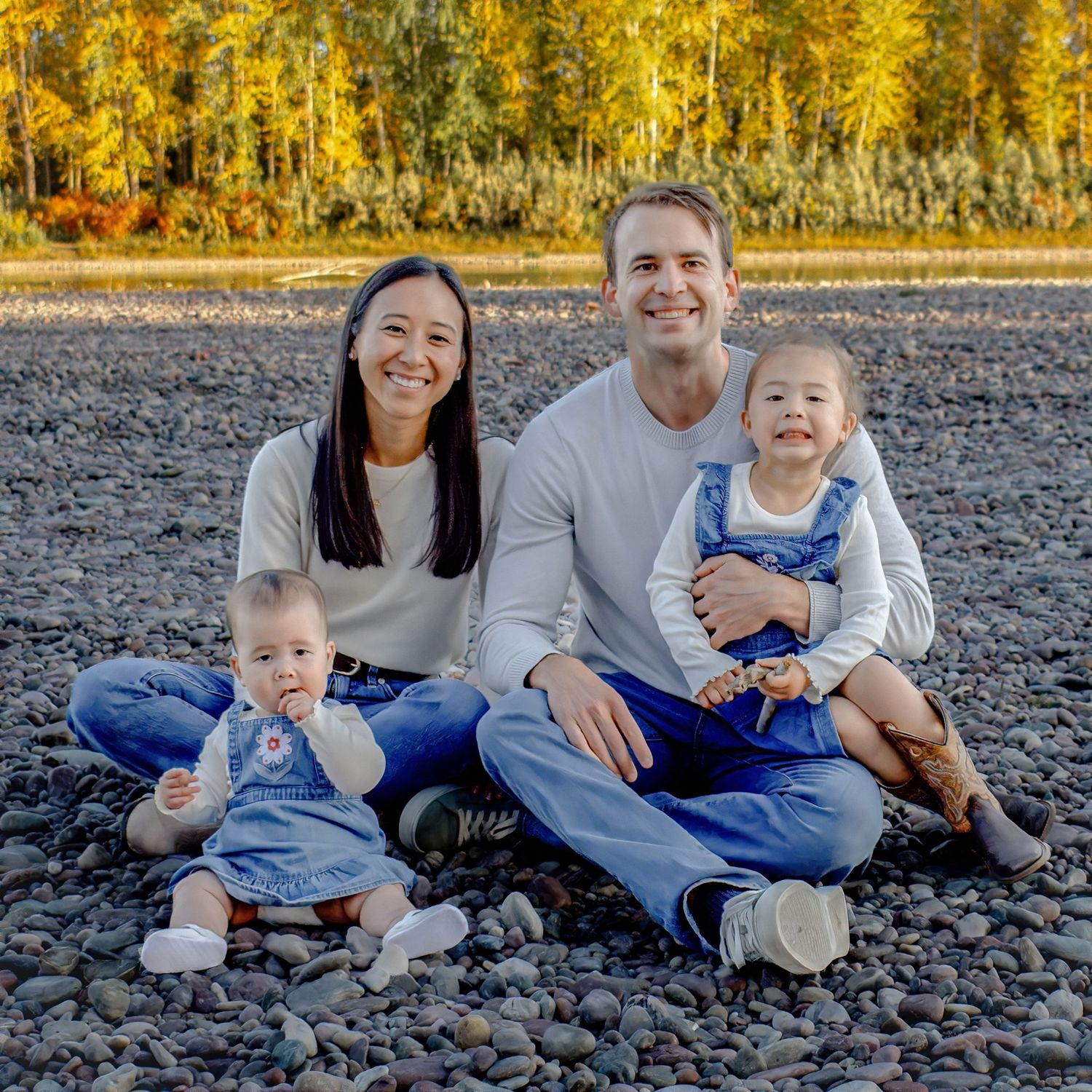 Family in coordinating blue and grey outfits sitting together on rocky beach during autumn sunset.