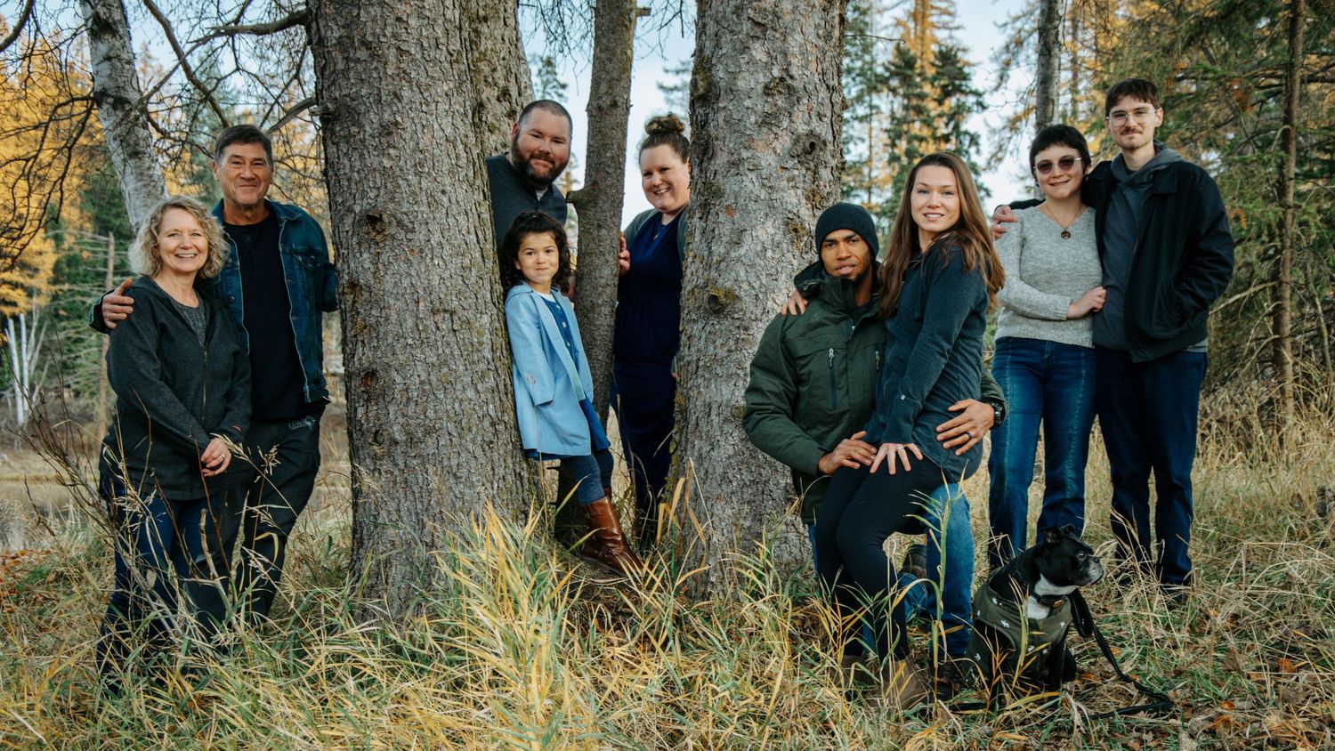 A group of people pose casually against trees in a wooded outdoor setting during autumn.
