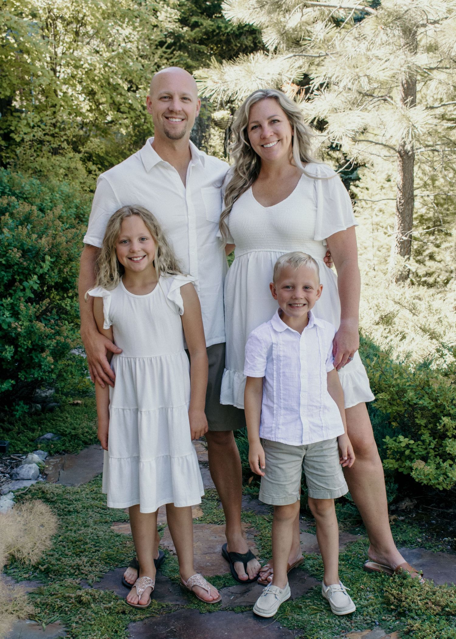 A family of four wearing coordinated white outfits poses together outdoors on a sunny day in a garden setting.