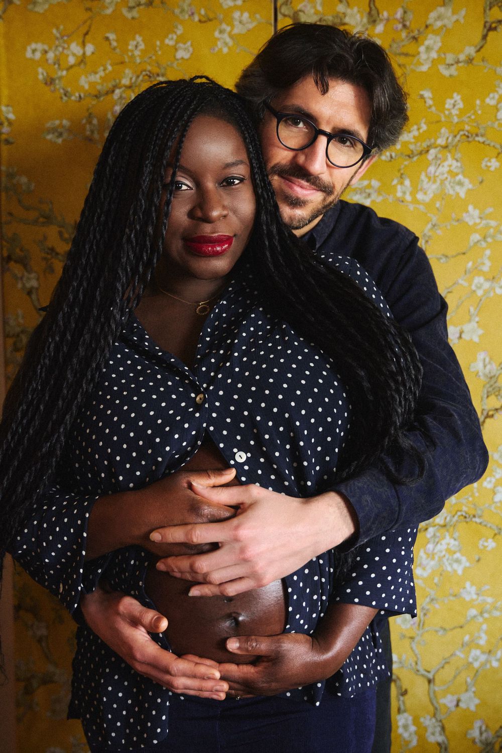 A couple poses for a pregnancy photo against a yellow textured wall wearing polka dot and navy outfits.