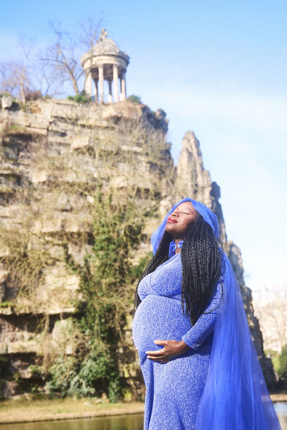 A pregnant woman in a flowing blue lace dress and veil poses at Buttes Chaumont park in Paris with Temple de la Sibylle visible.