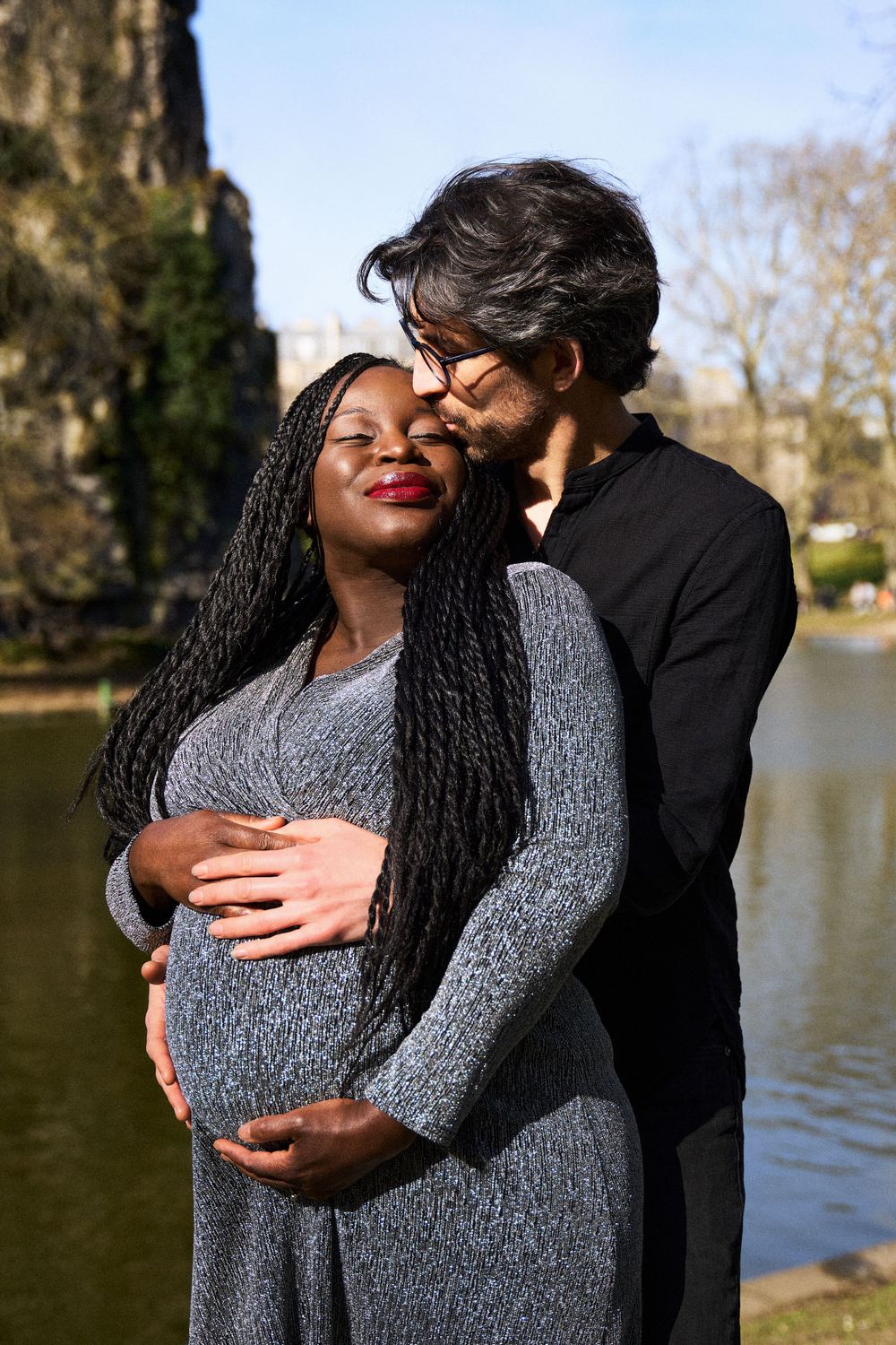 Romantic couple embraces by a lake on a sunny day, sharing an intimate moment while cradling an expectant belly.