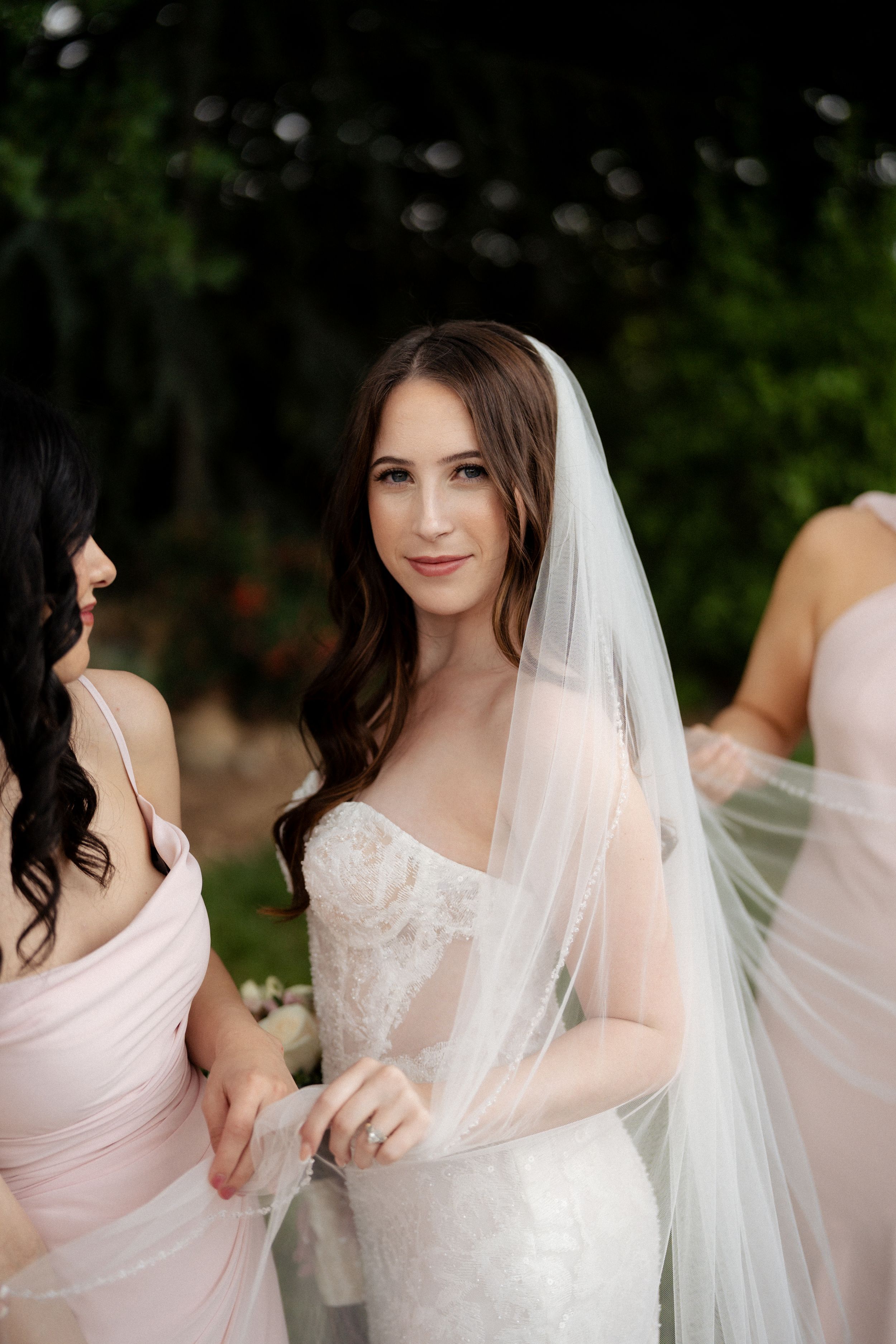 Smiling bride in lace gown and veil stands with bridesmaids in blush dresses. Fresno California Wedding Photographer and Videographer.