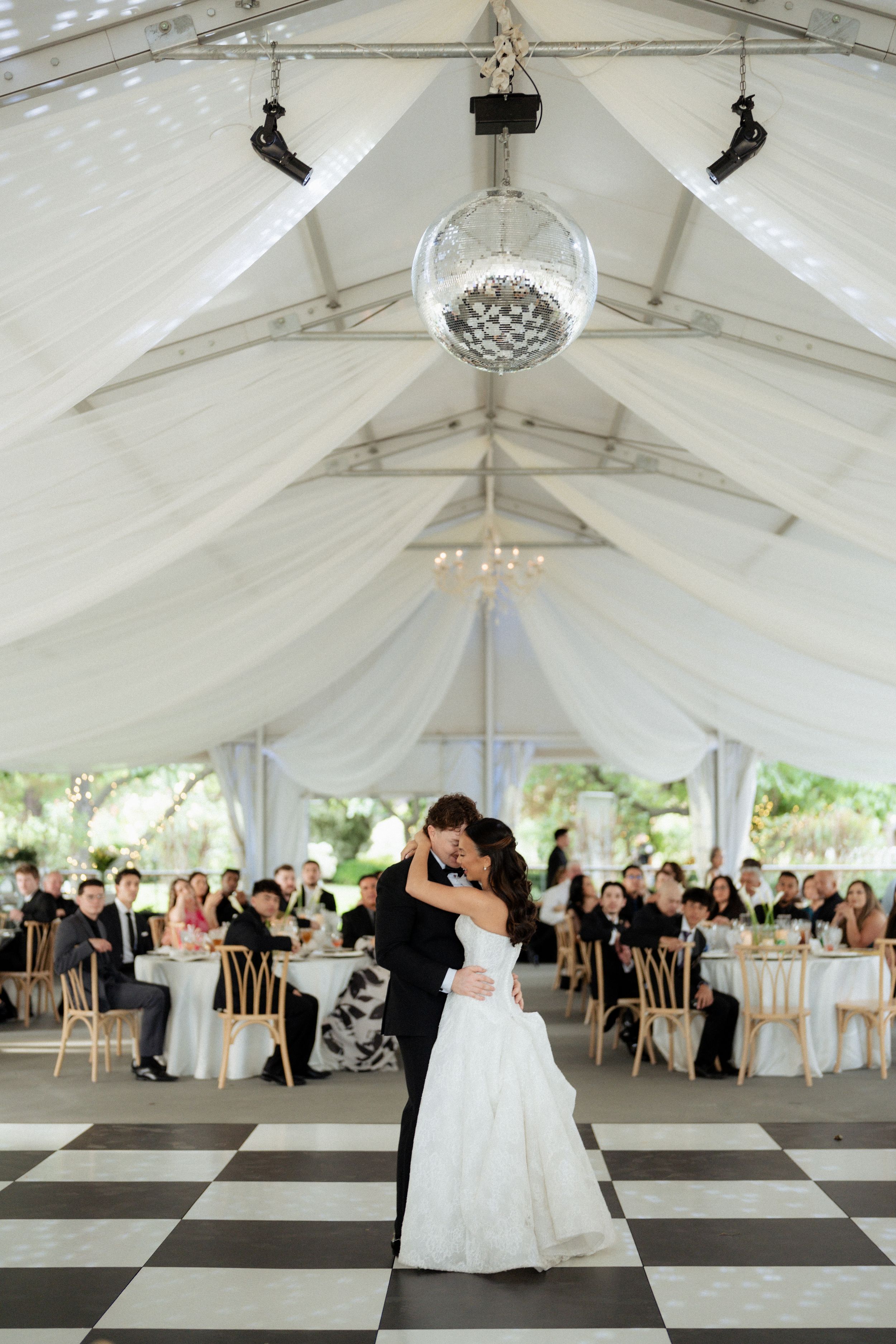 Bride and groom share first dance under disco ball, white draped tent with checkered dance floor. Fresno California Wedding Photographer