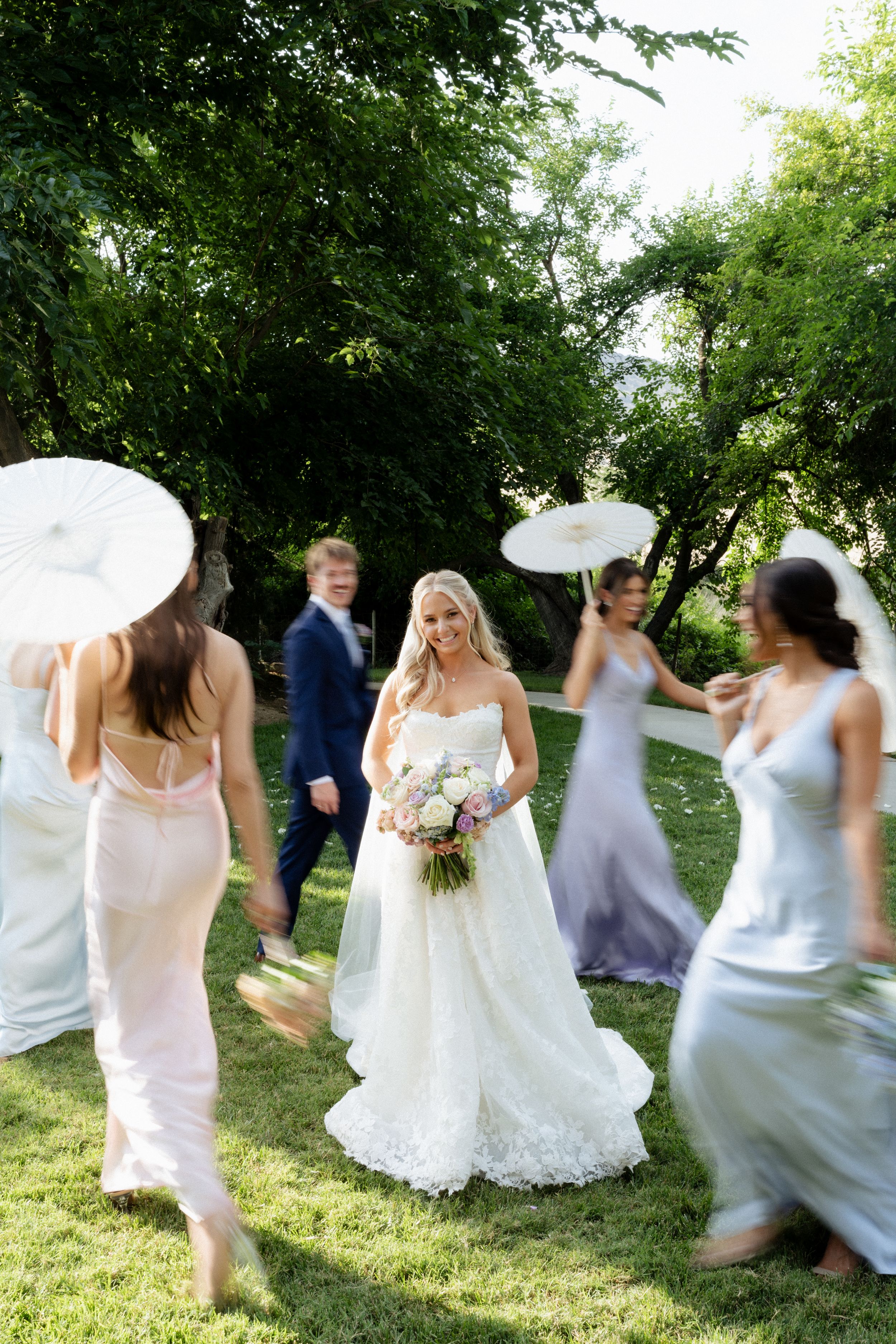 Blonde bride in lace gown holds bouquet surrounded by bridesmaids with parasols in sunny garden. Fresno California Wedding Photographer