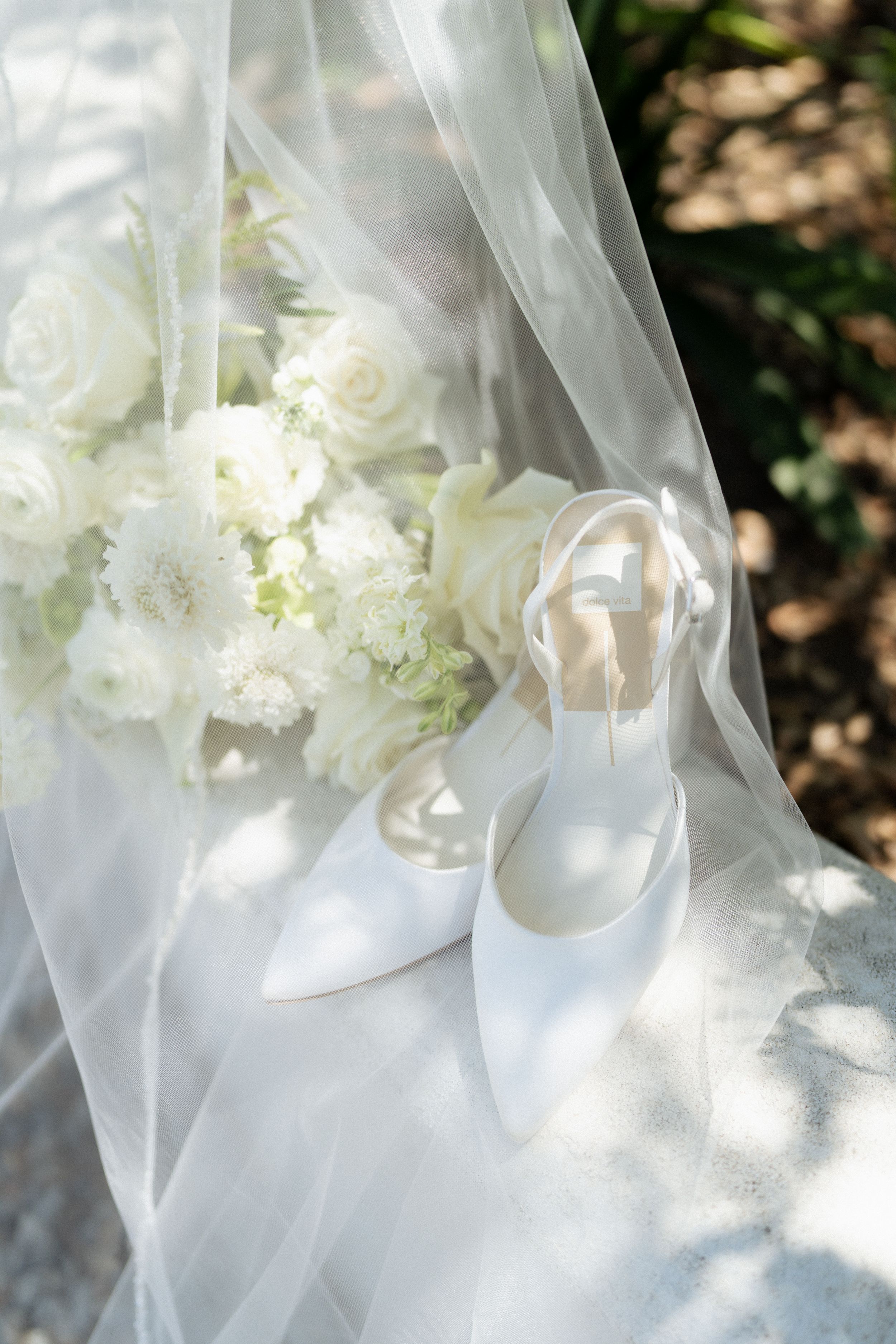 White bridal heels and white rose bouquet arranged under sheer veil in dappled sunlight outdoors. Fresno California Wedding Photographer