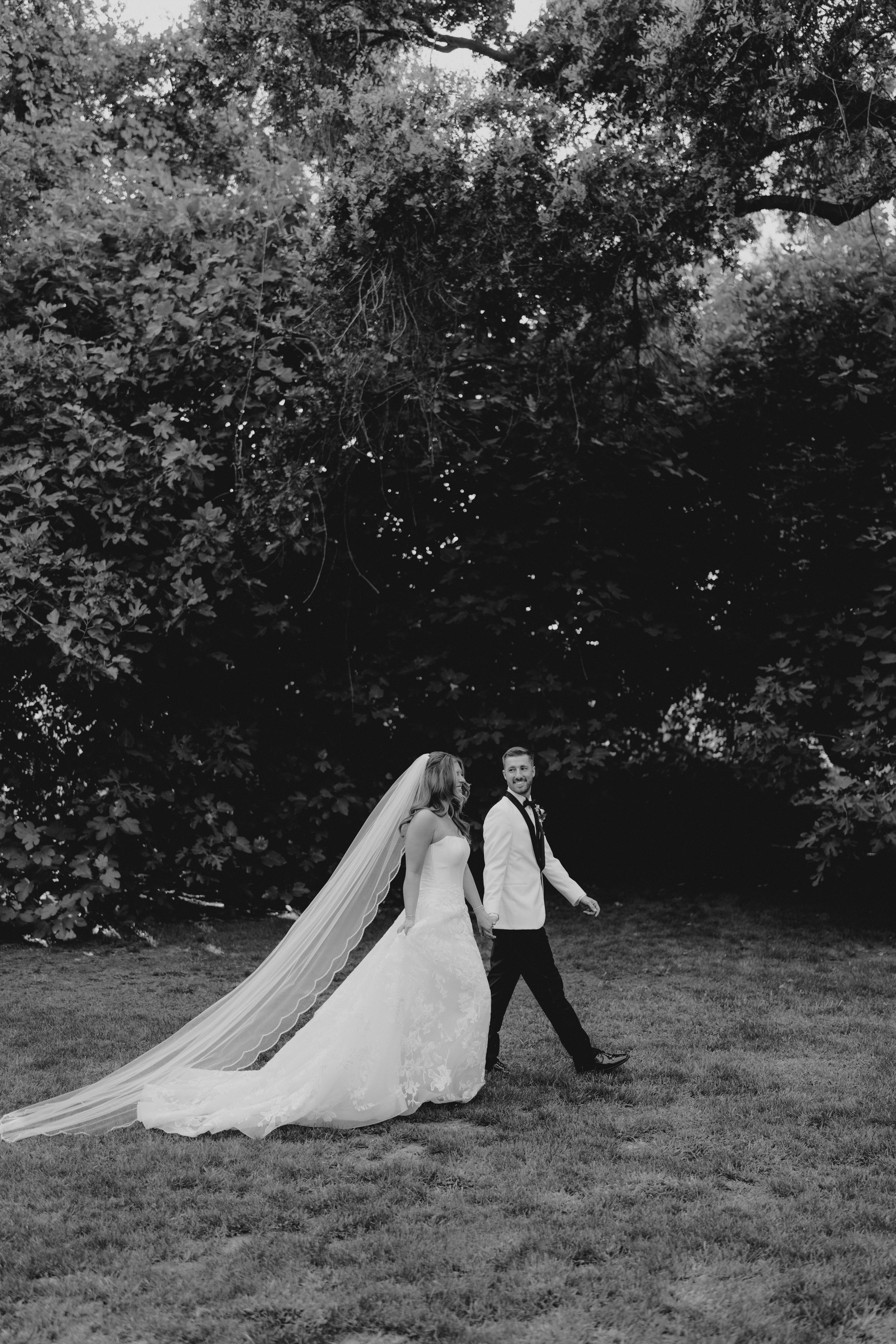 Black and white photo of bride and groom walking, long veil trailing behind. Fresno California Wedding Photographer and Videographer