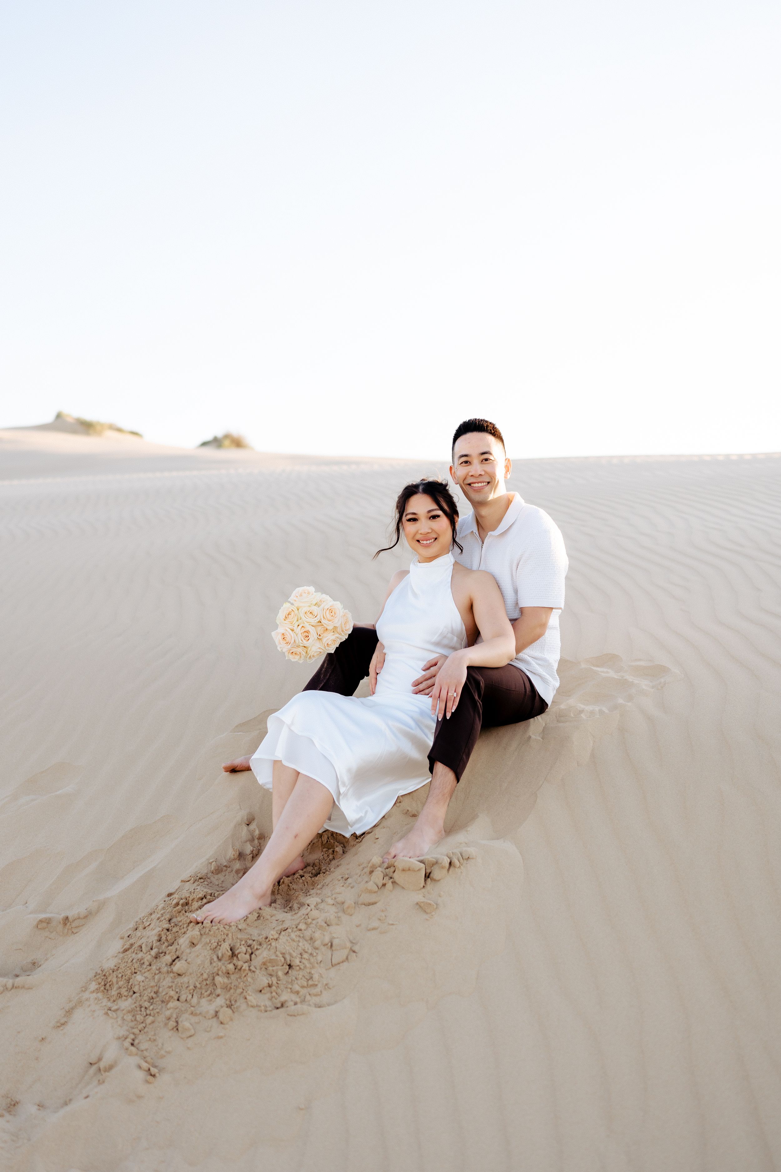 Couple sitting together on sandy desert dunes. Fresno California Wedding Photographer and Videographer