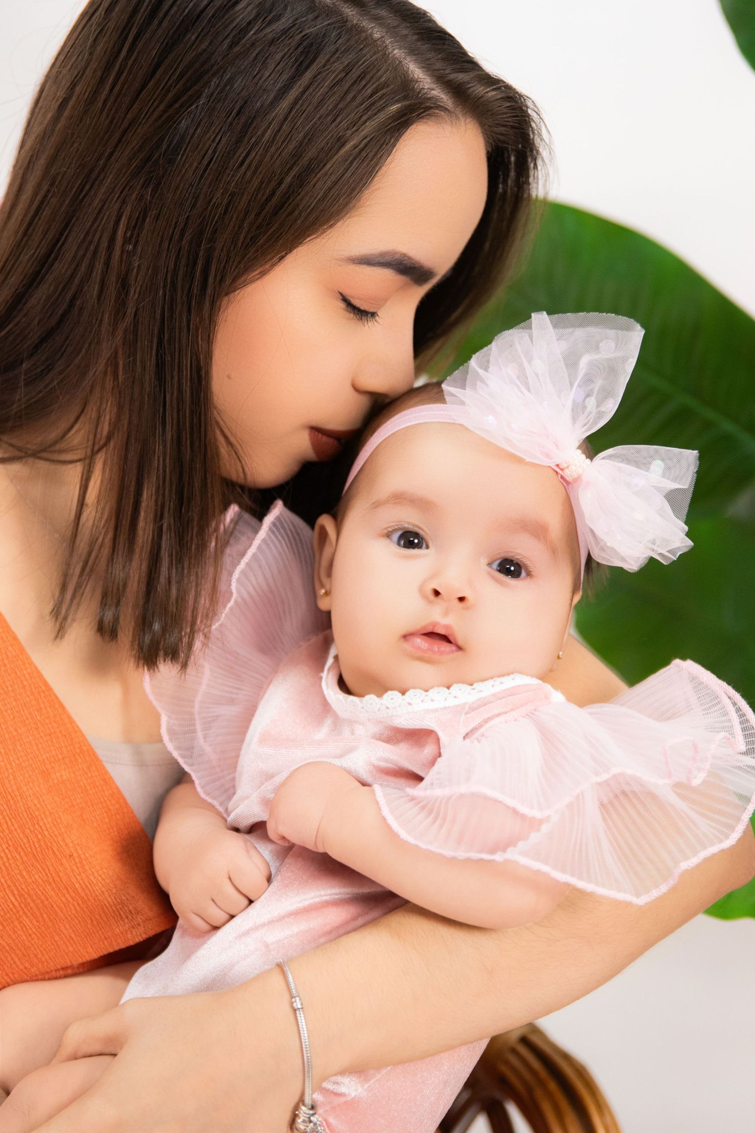 Young mother kissing her baby girl dressed in pink ruffled outfit with bow headband.