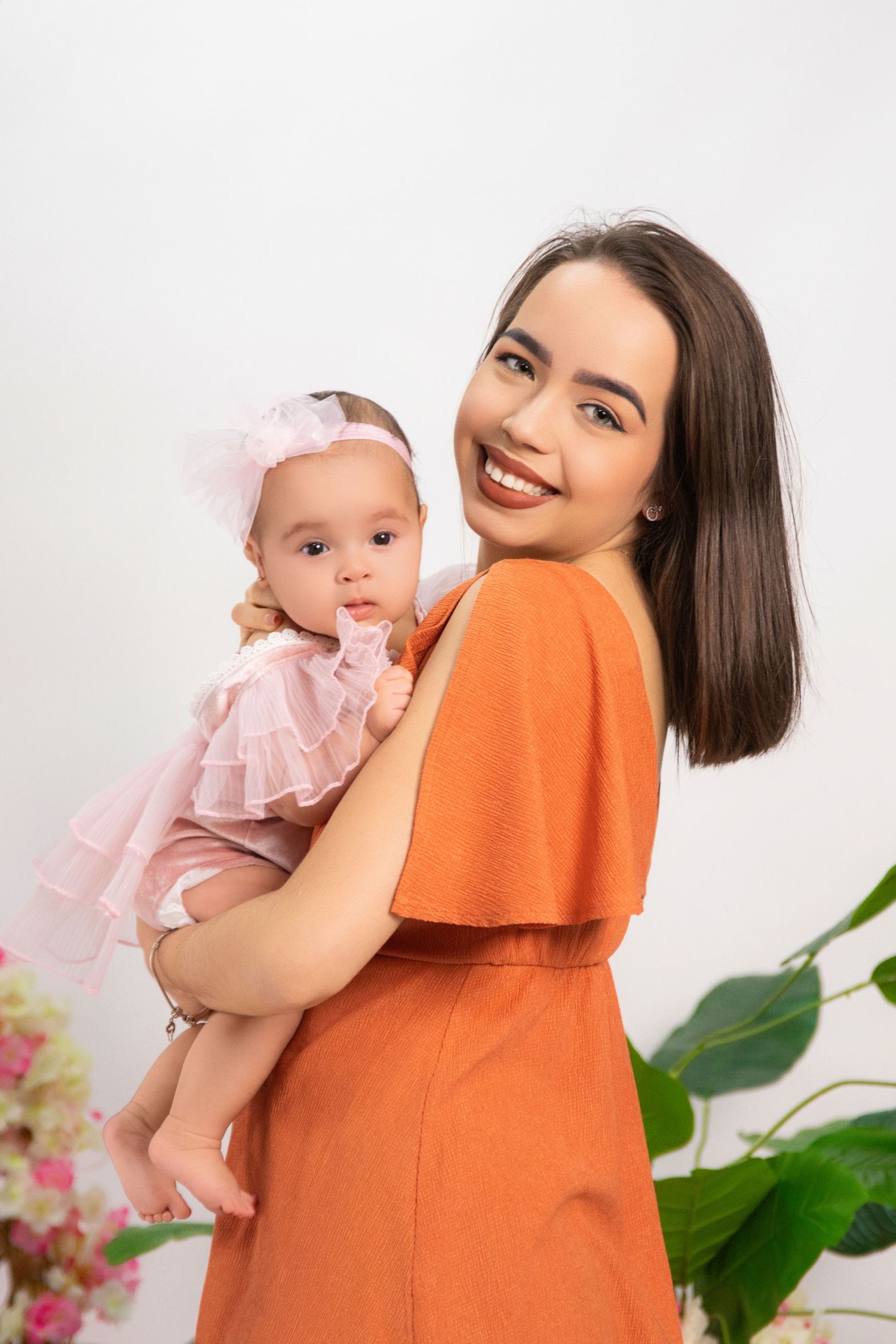 Smiling young mother in orange dress holding adorable baby girl in pink tutu dress and bow headband.