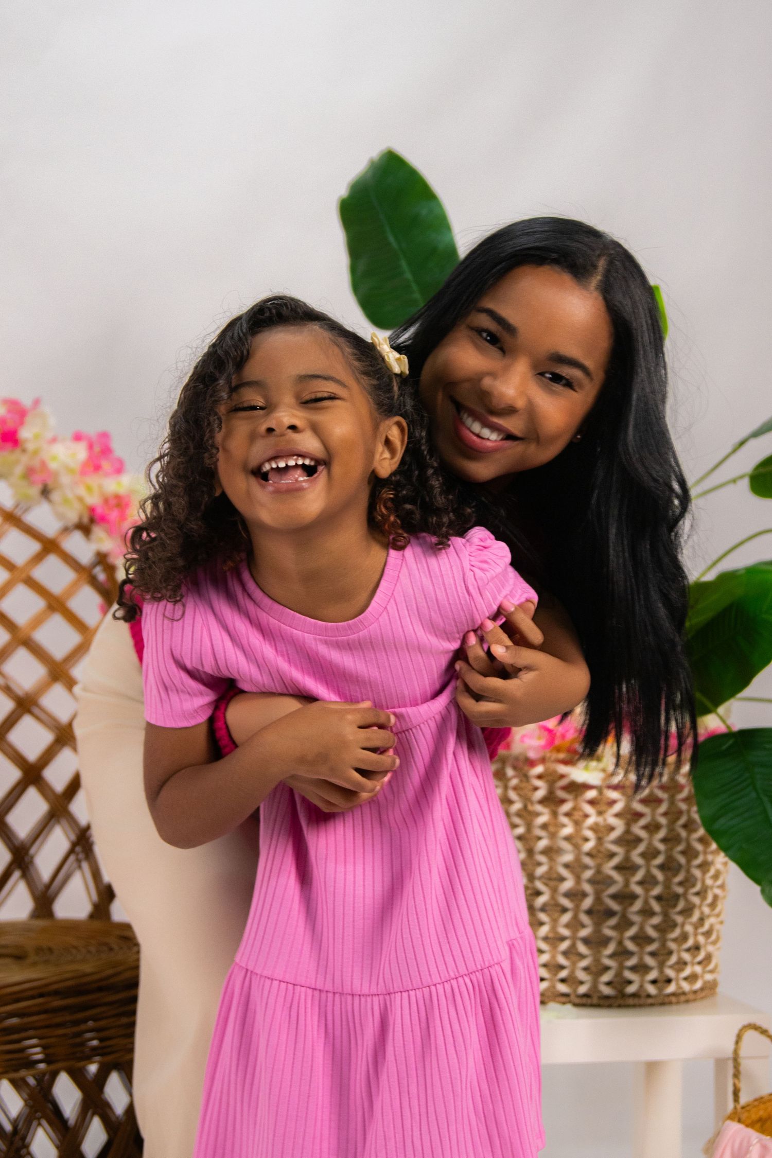 A joyful young Black girl in a pink dress laughs while an older girl hugs her from behind in a bright studio setting.