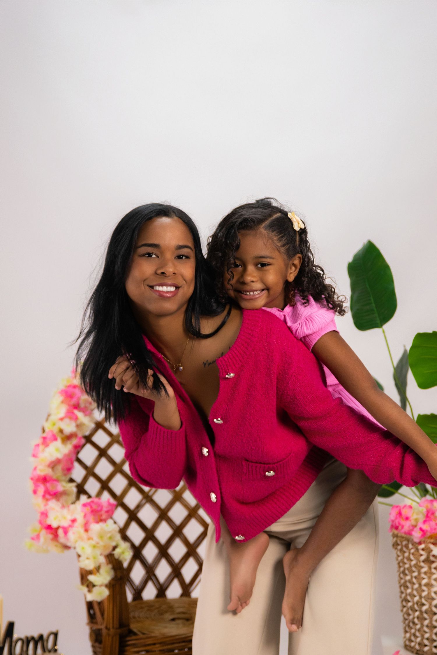 A woman and young girl in matching pink cardigans pose together, smiling, with floral decorations in the background.