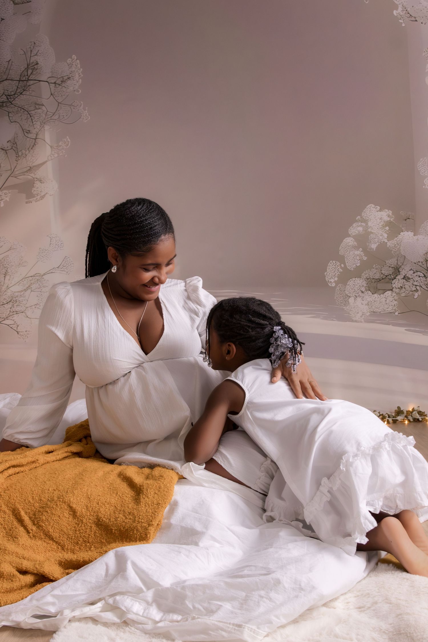 Pregnant Black woman in white dress sits with young daughter in matching white dress on studio floor with floral backdrop.