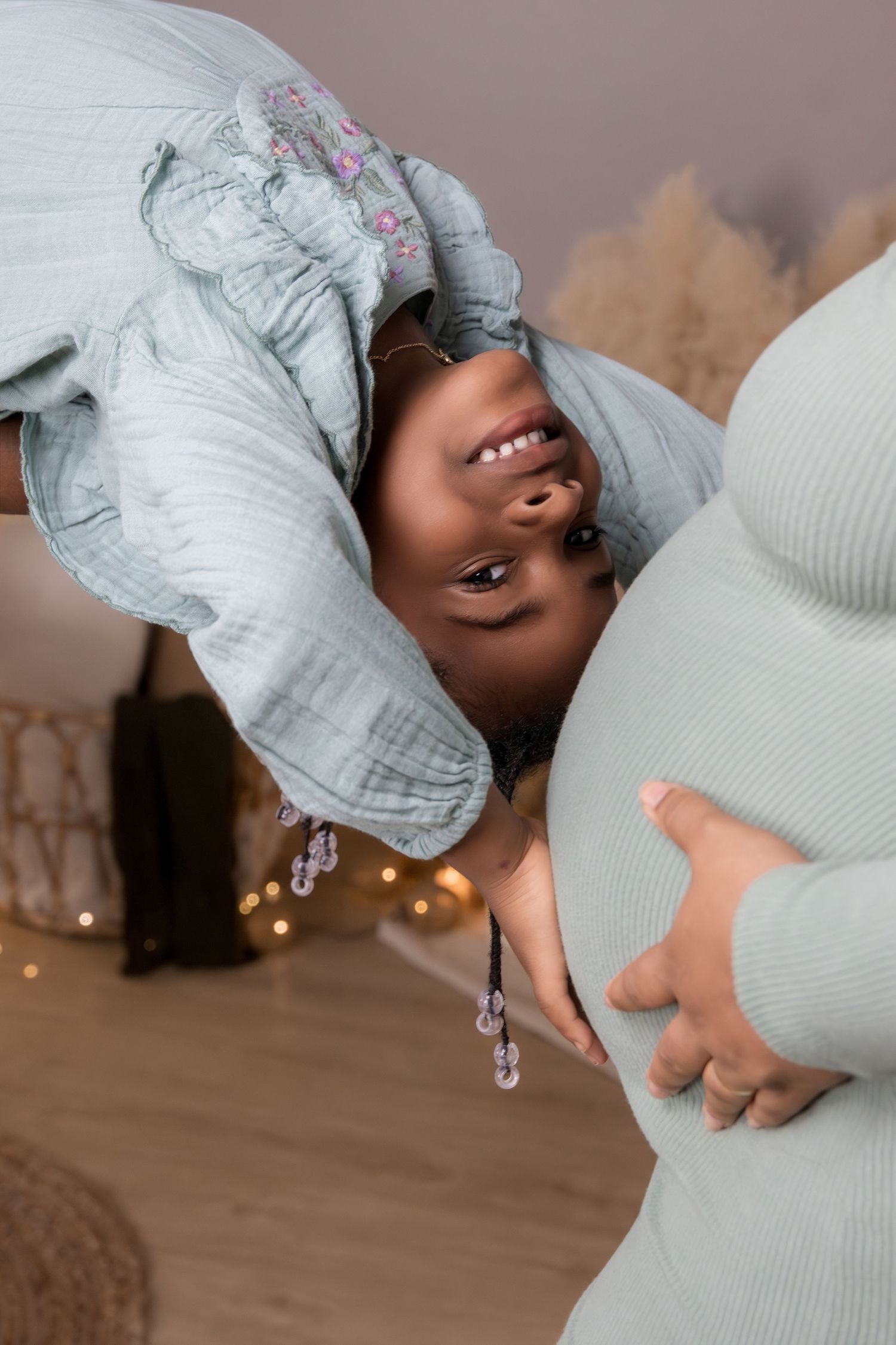 A young girl in a light blue outfit leans upside down, smiling, next to a pregnant woman's belly.