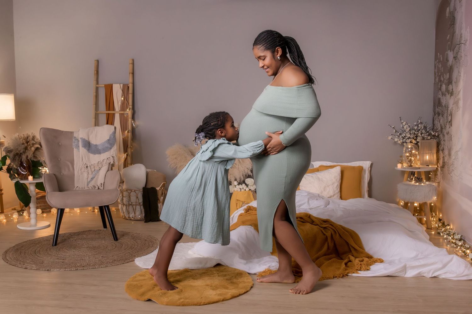 Pregnant Black woman in green dress dancing with young daughter in cozy bedroom with warm holiday lights.