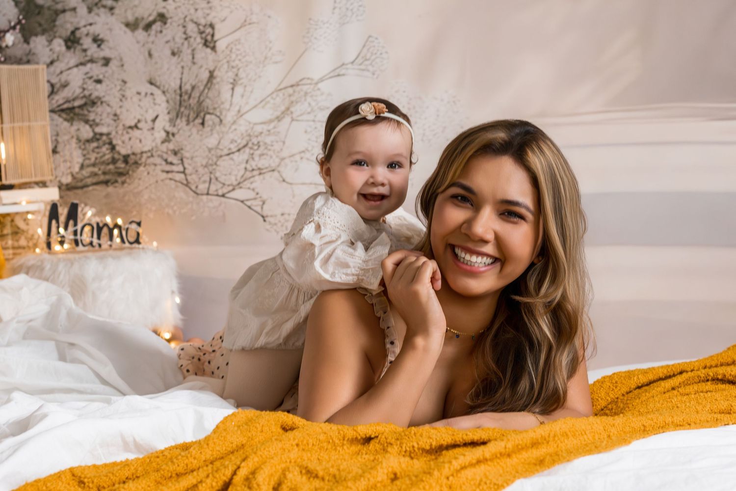 Smiling mother lying on bed holding happy baby girl, yellow blanket, white decor background.