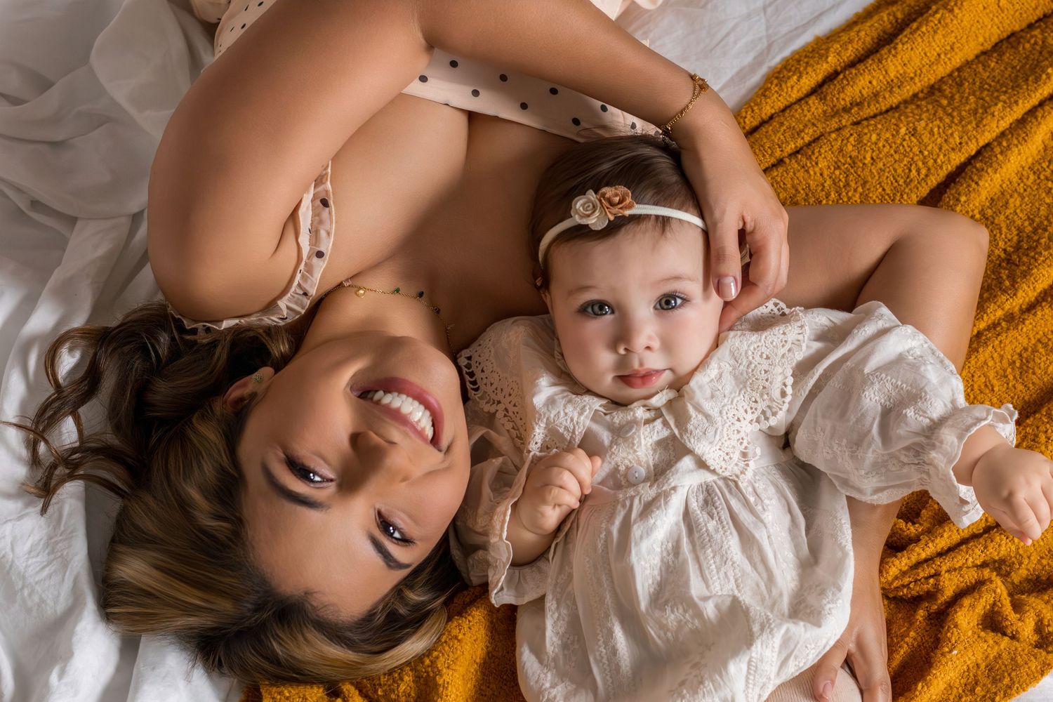 A smiling mother and her baby girl in white dress lying together on a golden yellow blanket, viewed from above.