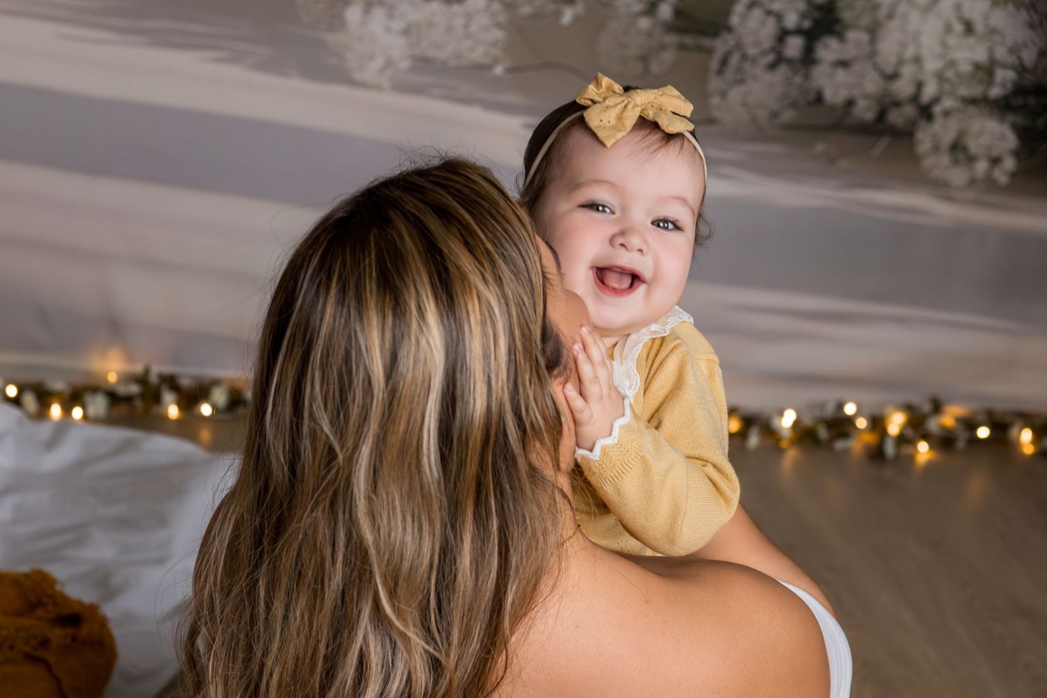A smiling baby girl in a yellow outfit and bow headband being held by her mother indoors with warm fairy lights.