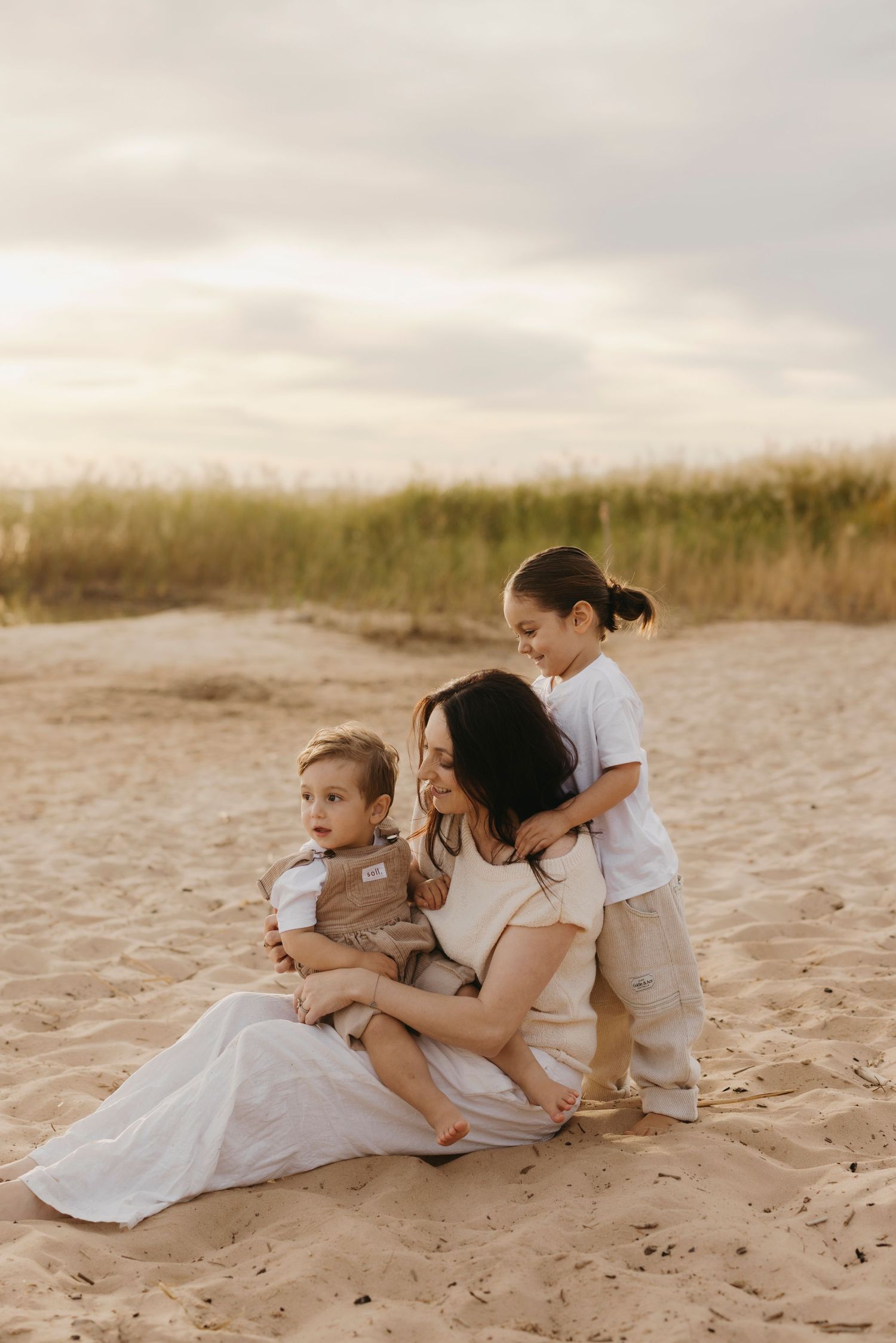 Mother sitting on beach sand with two young children during golden sunset hour.