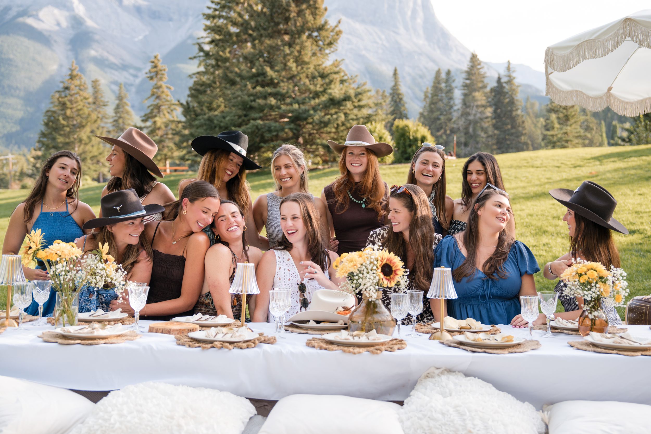 Group of women in hats celebrating at an outdoor table with mountain views and sunflower arrangements.