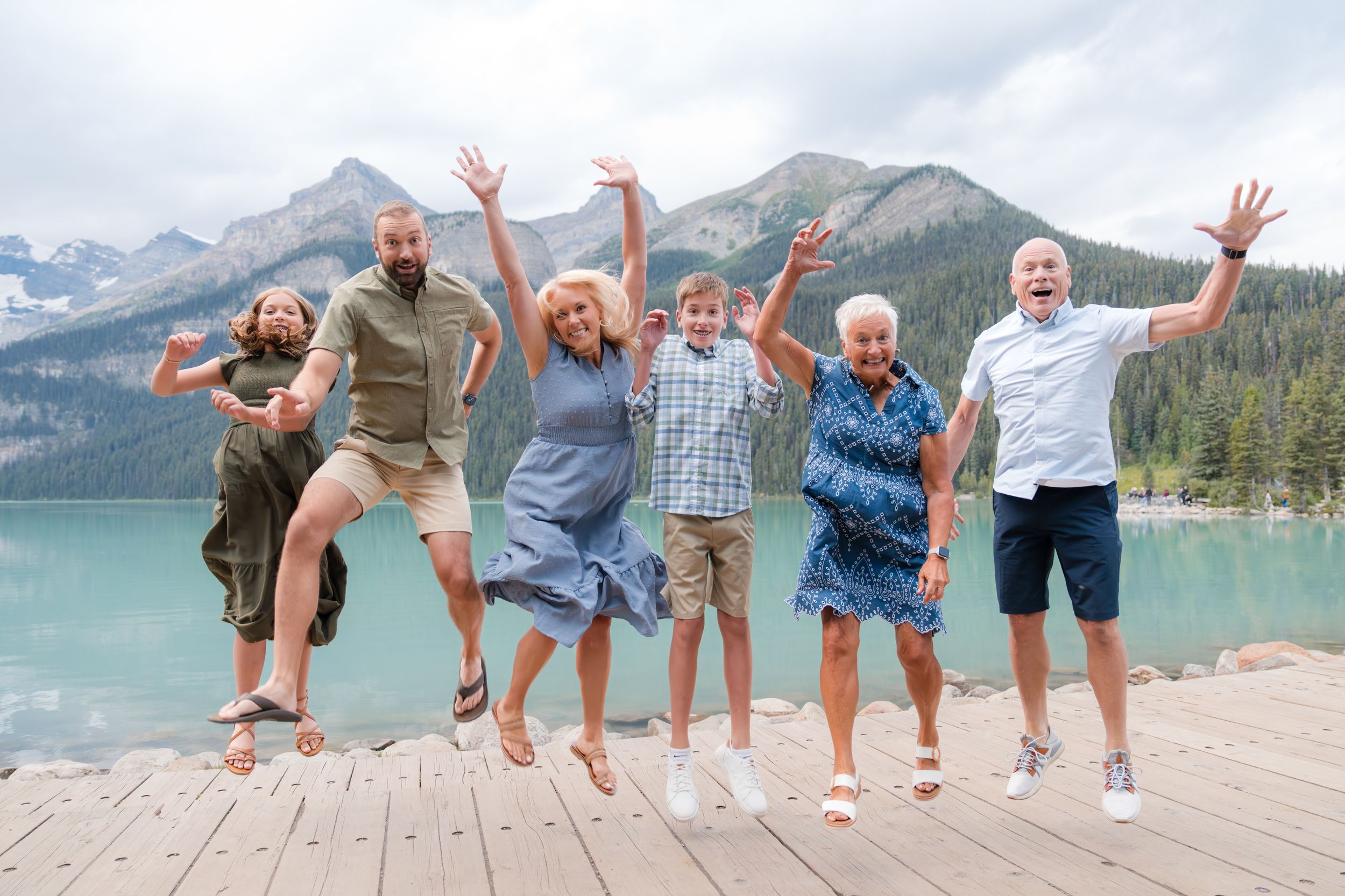 Six people jumping joyfully on a wooden dock with a turquoise mountain lake and snowy peaks in the background.