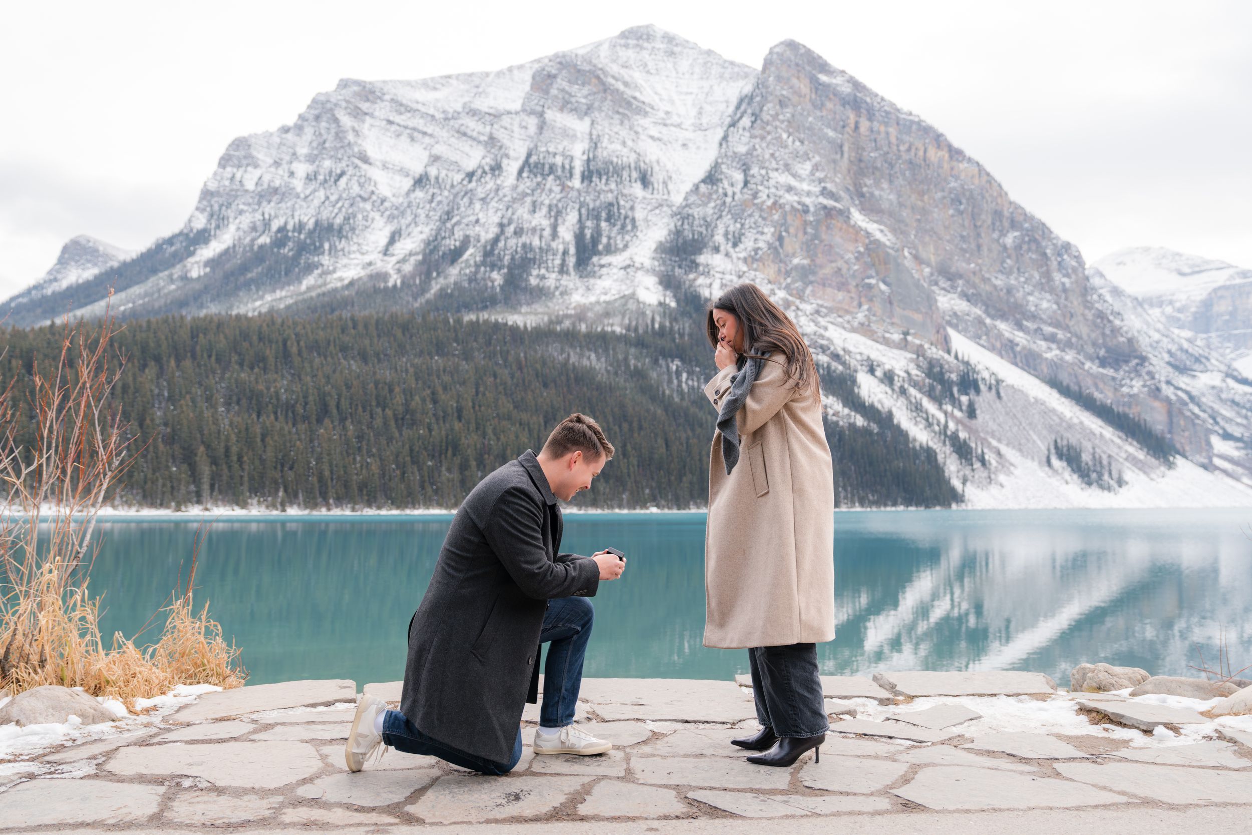 Man proposes on one knee to woman at Lake Louise with turquoise water and snowy mountains backdrop.