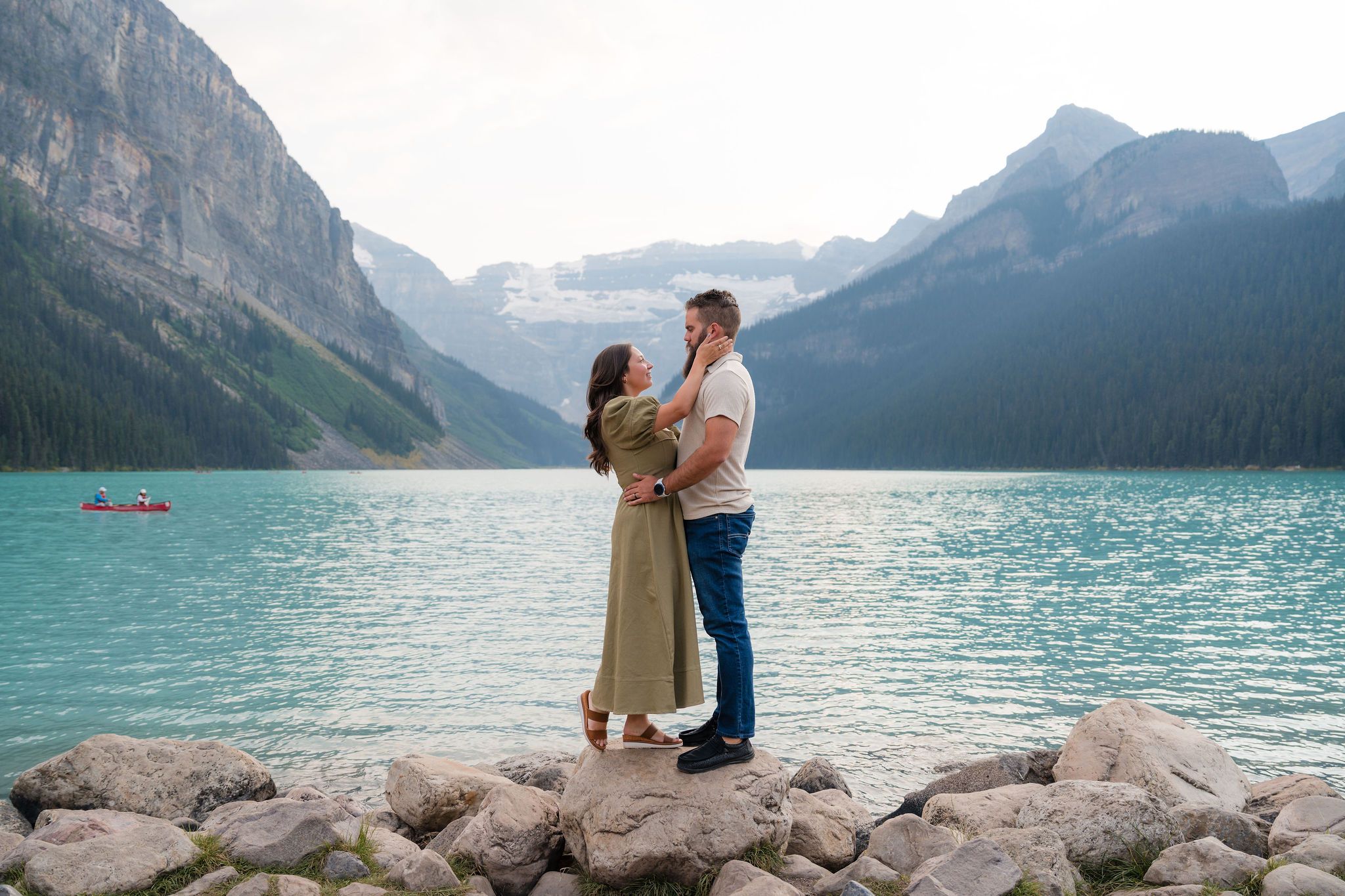 Couple embracing romantically on rocky shore of turquoise Lake Louise with majestic Canadian Rockies mountains in background.