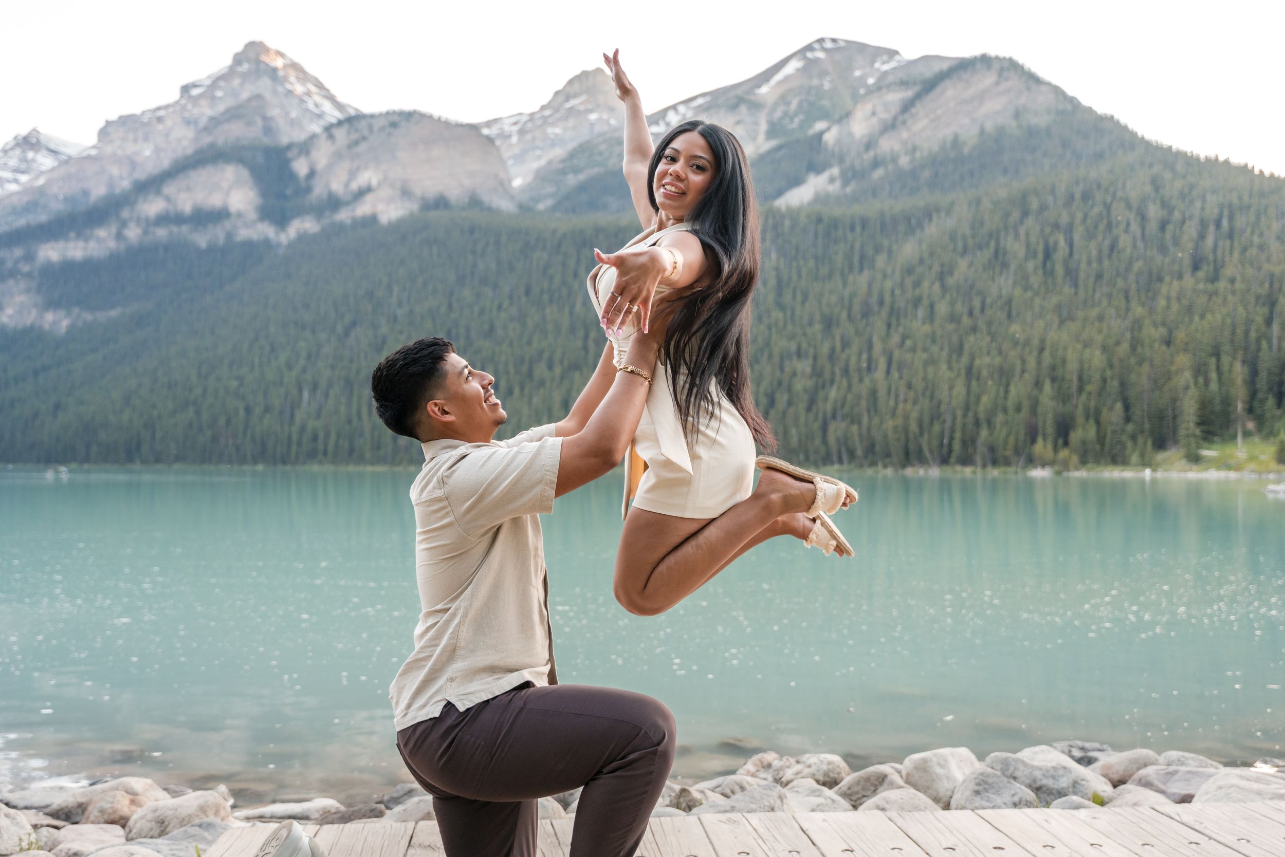 Woman jumps in air showing off engagment ring as fiance looks up at her