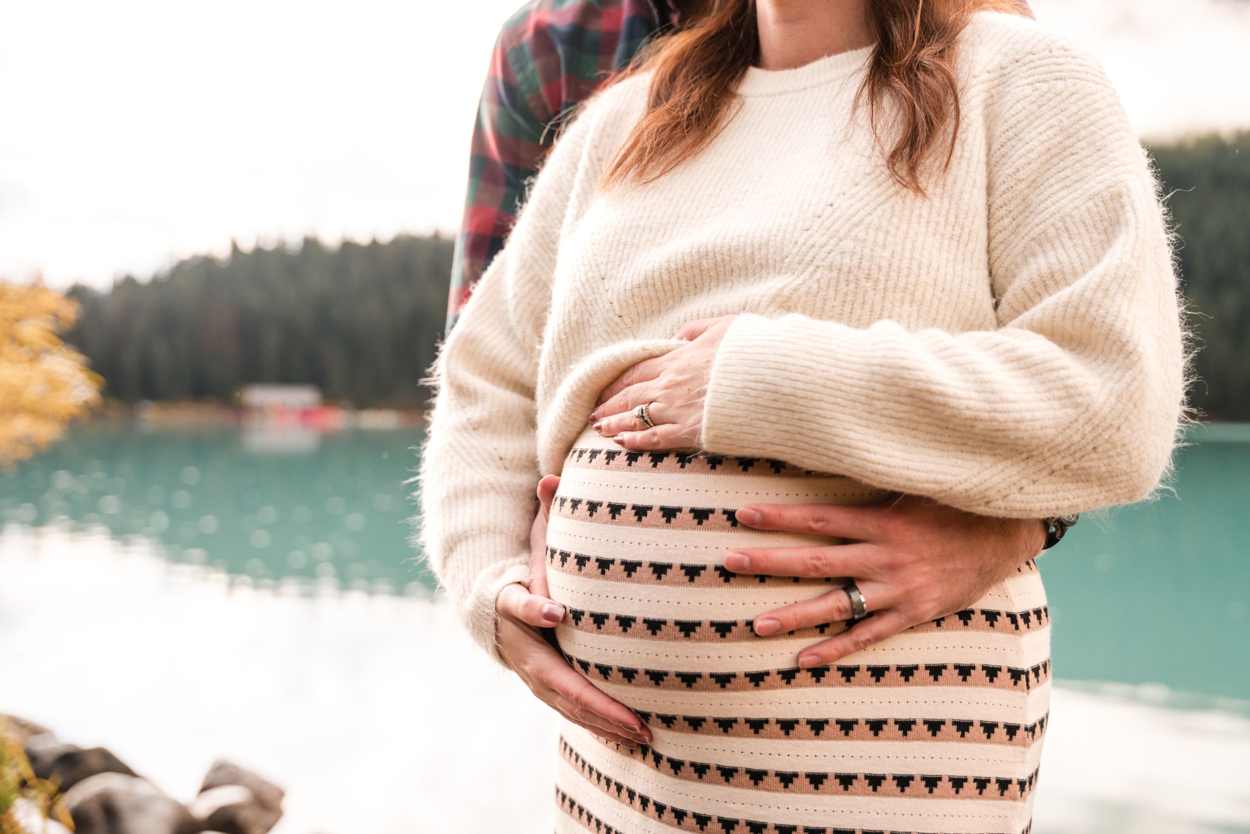 Close-up view of a pregnant belly in a cream colored sweater with triangle pattern against a lakeside backdrop.