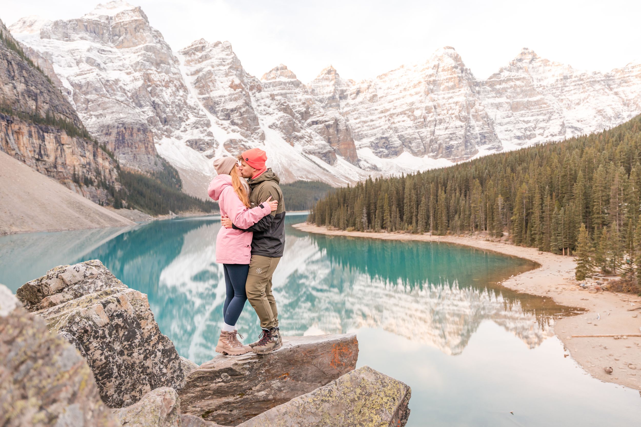 Couple embracing on rocks at Moraine Lake, Banff, with turquoise water and snow-capped Rocky Mountains reflected behind them.