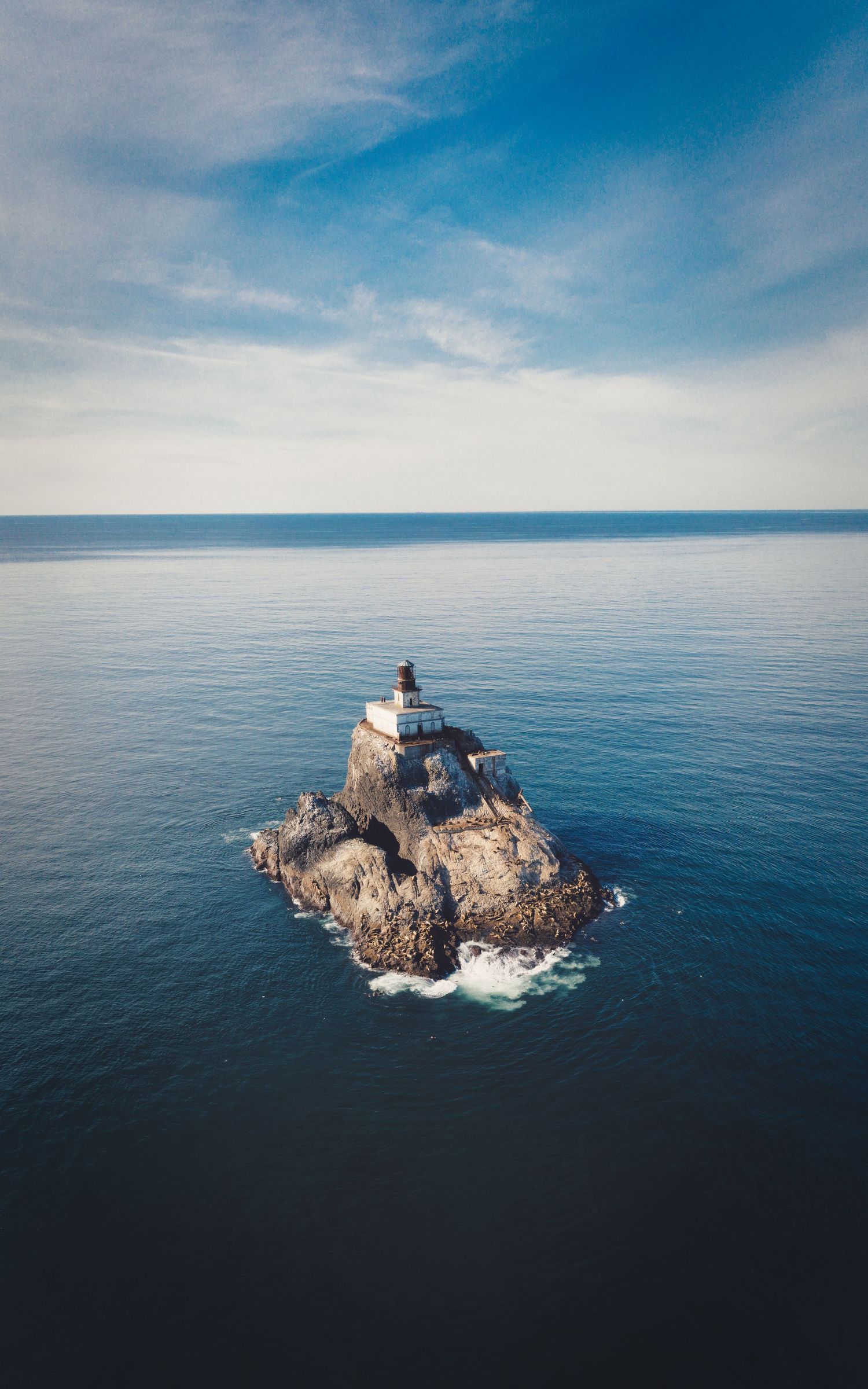 Aerial view of a solitary lighthouse perched atop a rocky island outcrop in the vast blue ocean under clear skies.