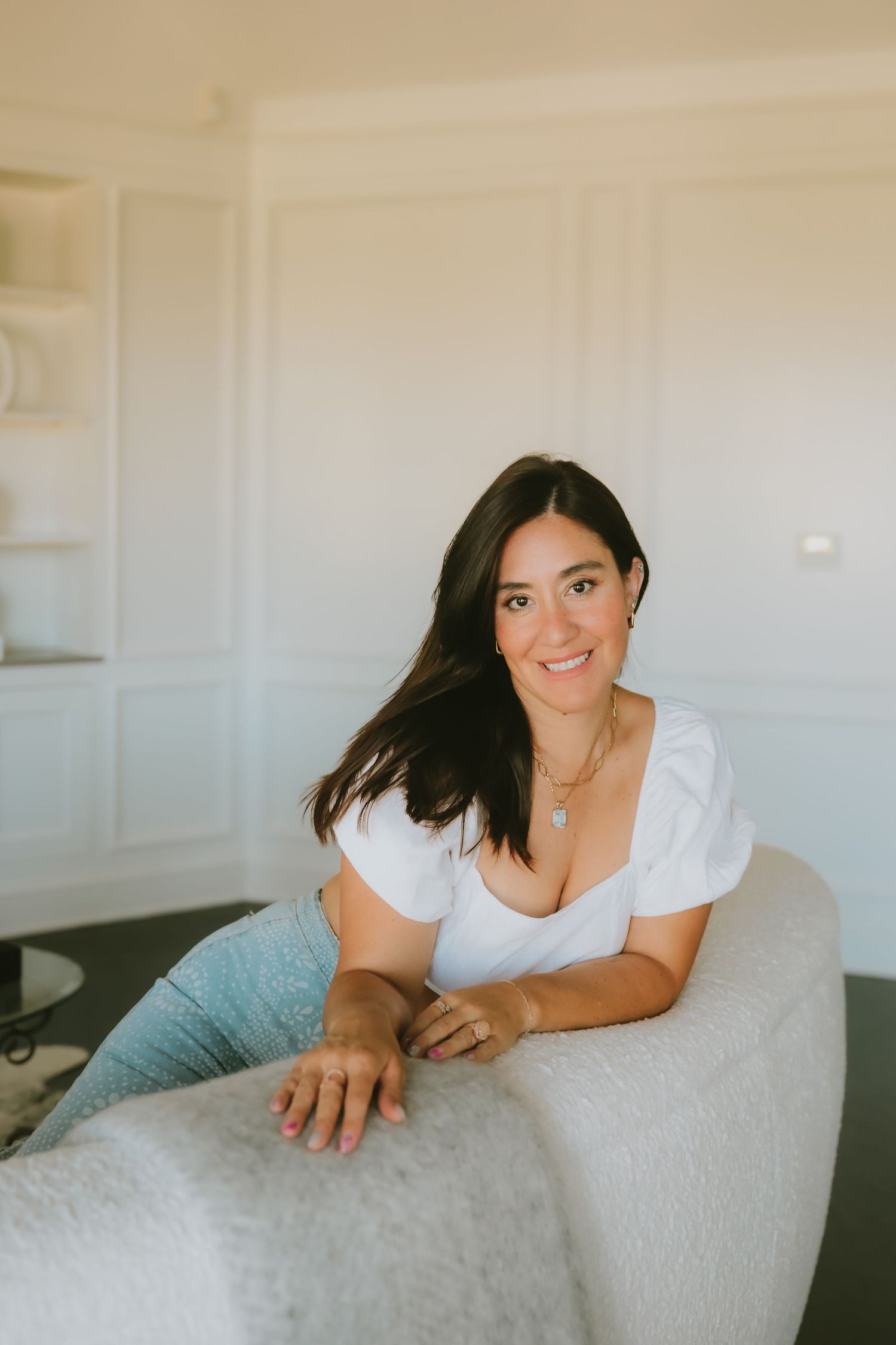 A smiling woman in a white top relaxes on a light gray armchair in a bright, minimalist interior setting.