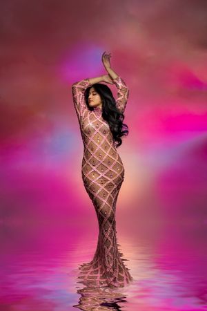 Woman in beaded pink gown poses gracefully in water with arms raised against a vibrant pink cloudy backdrop.