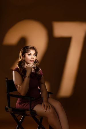 A young woman in a burgundy dress sits in a director's chair, posing elegantly in front of a glowing number 27 backdrop.