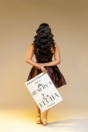 Girl in a satin brown dress with long curly hair, seen from behind, holding a Reserva la Fecha quinceañera save-the-date sign.
