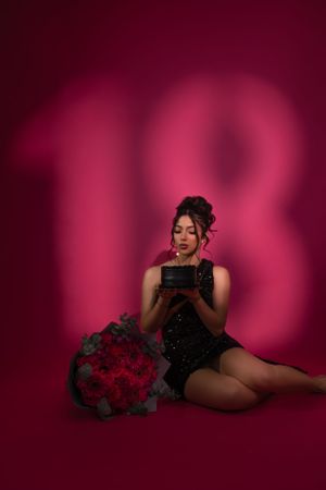 Figure in black dress poses against burgundy backdrop with number 18 projection and red roses.