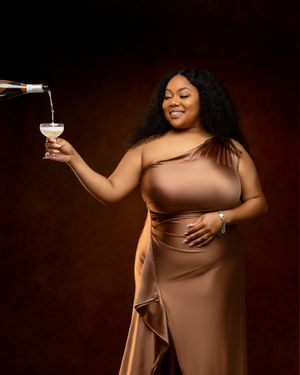 A figure in a brown dress poses elegantly with a wine glass against a dark background in a glamour photoshoot.