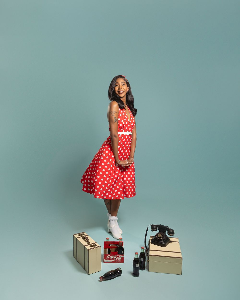 Young woman in red polka dot dress poses with vintage Coca-Cola items and rotary phone on blue background.