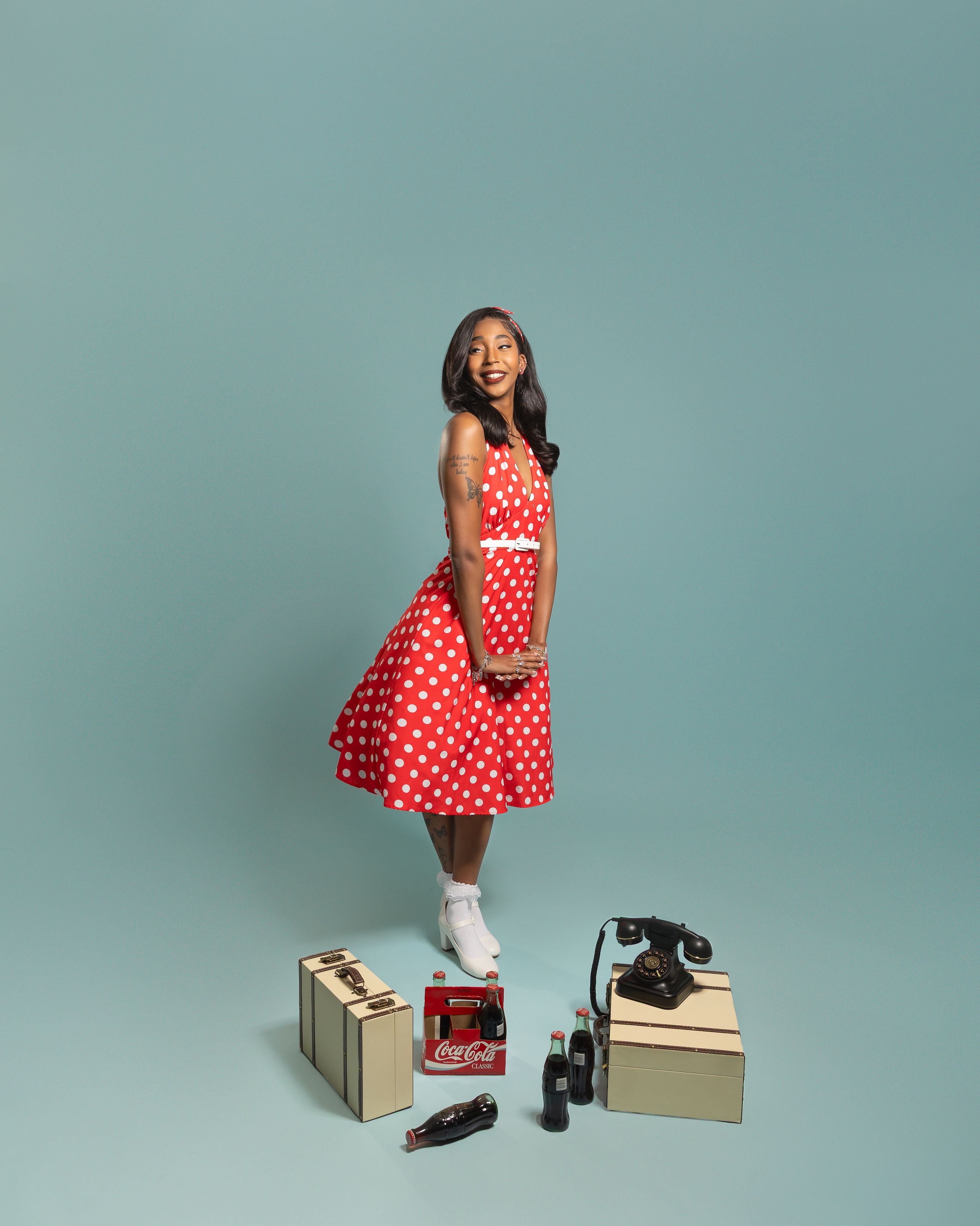 Young woman in red polka dot dress poses with vintage Coca-Cola items and rotary phone on blue background.