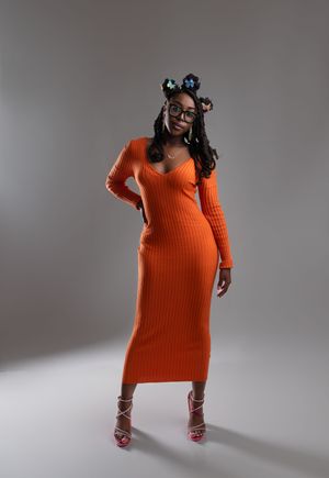 Long-sleeved orange ribbed bodycon maxi dress with red strapped heels and stylish hair accessories on grey backdrop.