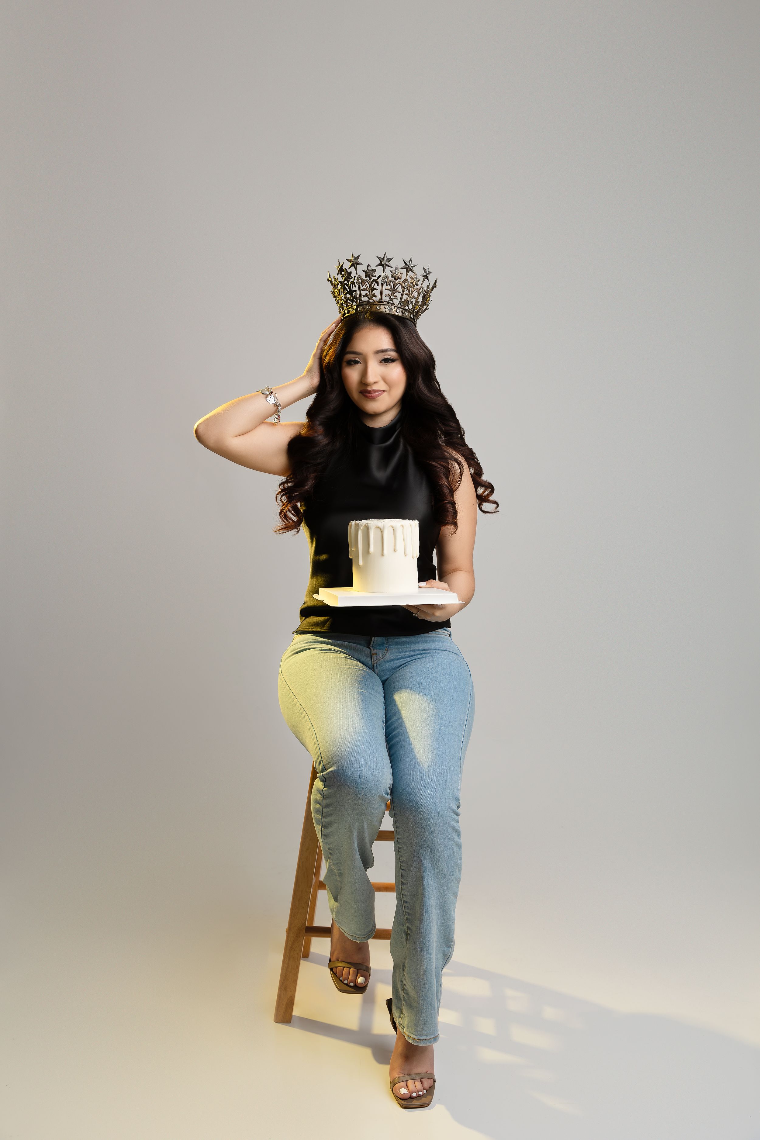 A figure wearing a crown and casual attire poses on a wooden stool against a light gray studio background.