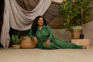 Someone wearing an elegant emerald green outfit reclines on the floor in a studio setting with draped fabric and potted plants.