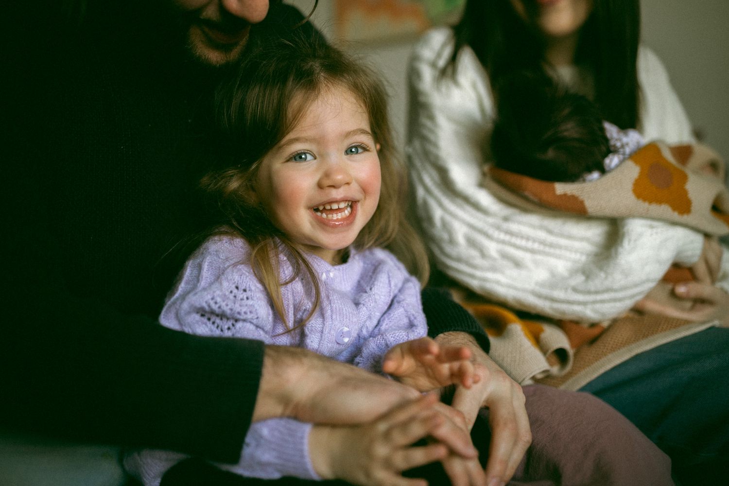 A family relaxes together on a porch couch of a white-sided house on a sunny afternoon.