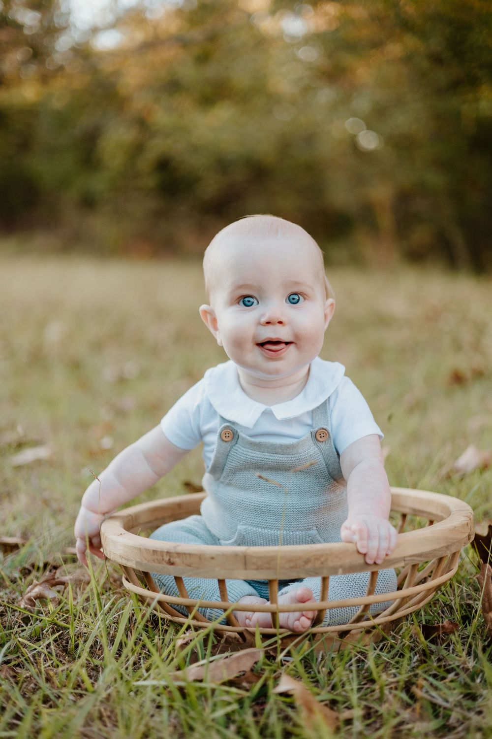 Happy baby sitting in wicker basket outdoors wearing light blue outfit and overalls.
