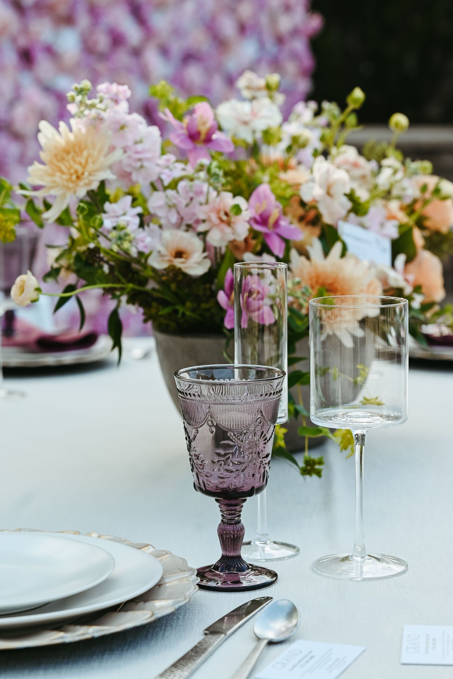 Elegant table setting with purple vintage glassware and fresh floral centerpiece featuring peach and pink blooms.