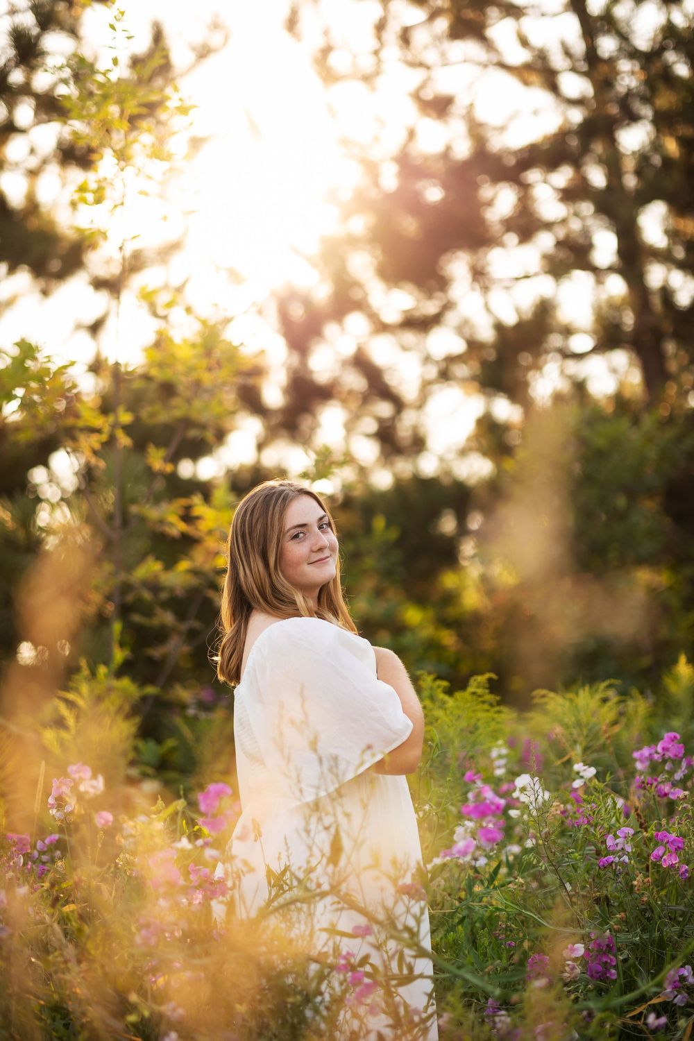 Dreamy golden hour portrait in white dress among wildflowers and trees.