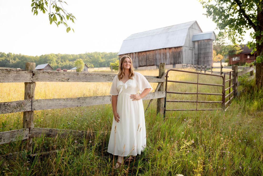 Rustic countryside portrait in white dress near weathered barn and wooden fence.