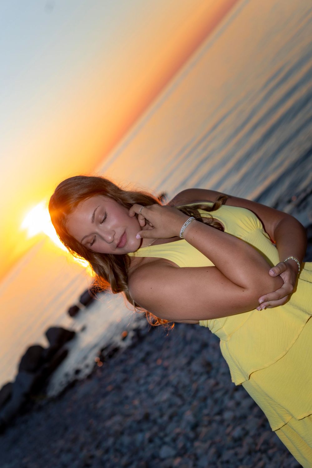 Woman in yellow dress posing on rocky beach at sunset with golden light reflecting on calm ocean water.